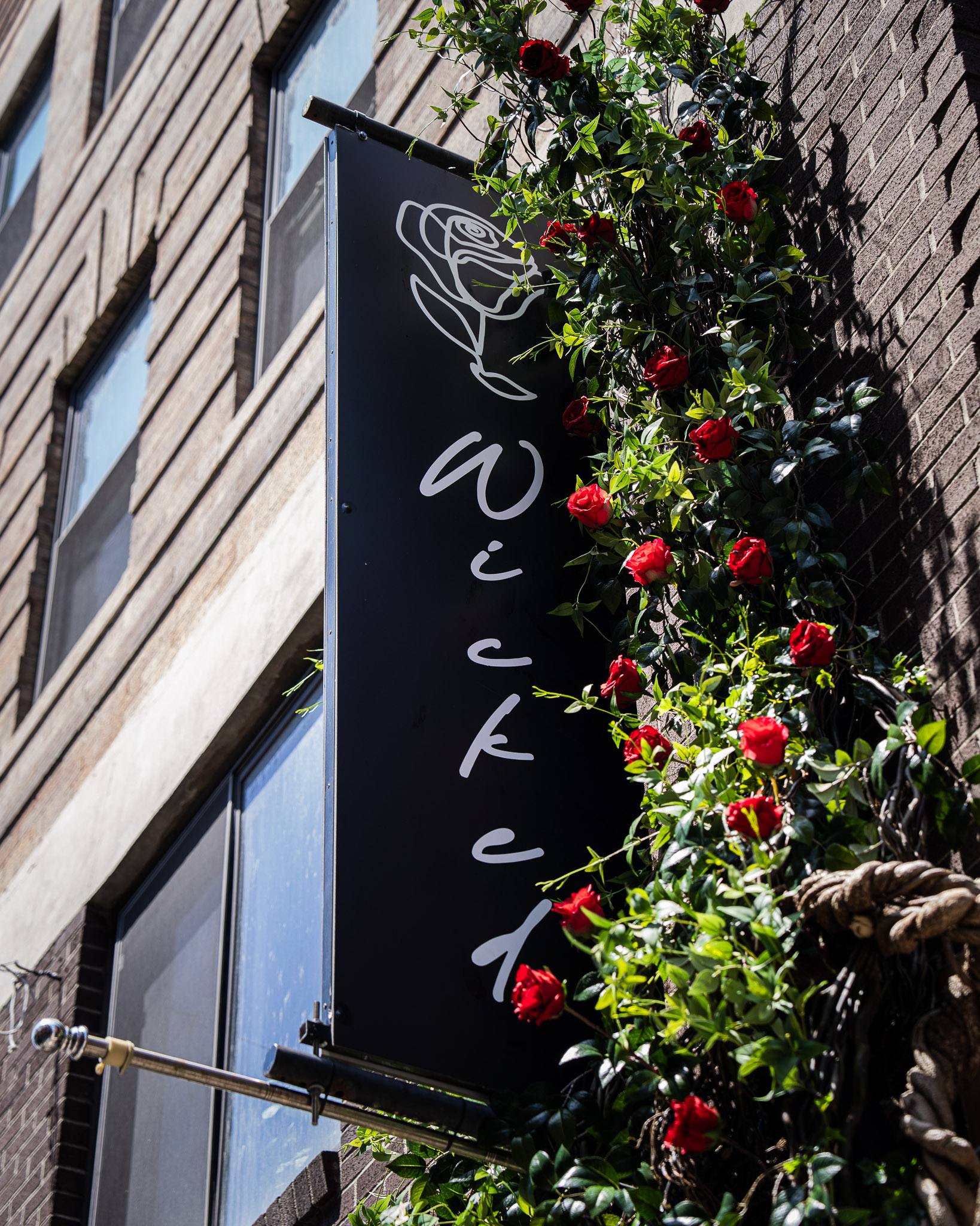 The black blade sign with a white line-drawn rose and the Wicked Craft Co. script, climbing red roses on dark brick.