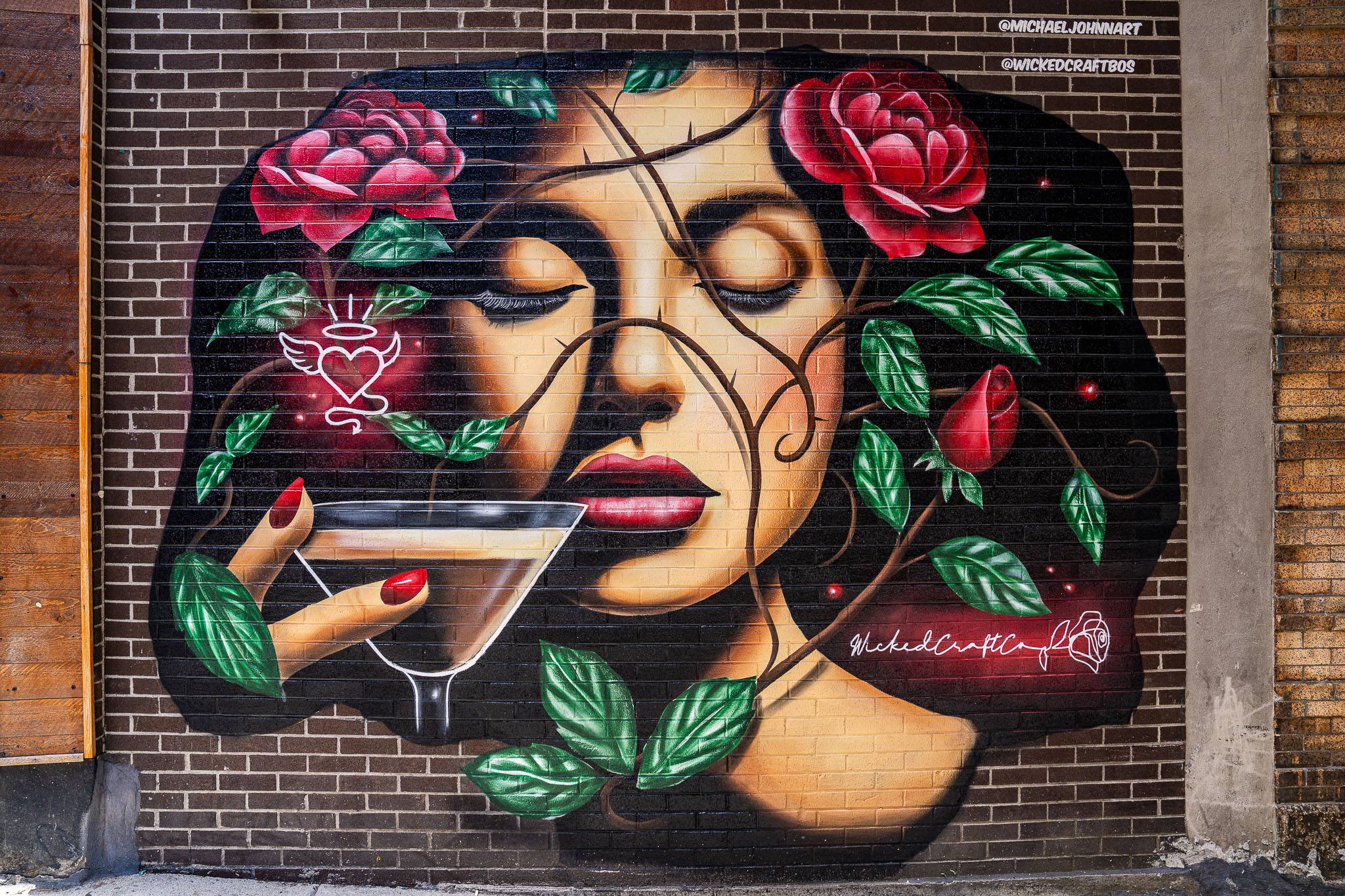 A painted mural of a woman in a wide-brimmed hat with roses and a martini, signed @michaeljohnnart and @wickedcraftbos.