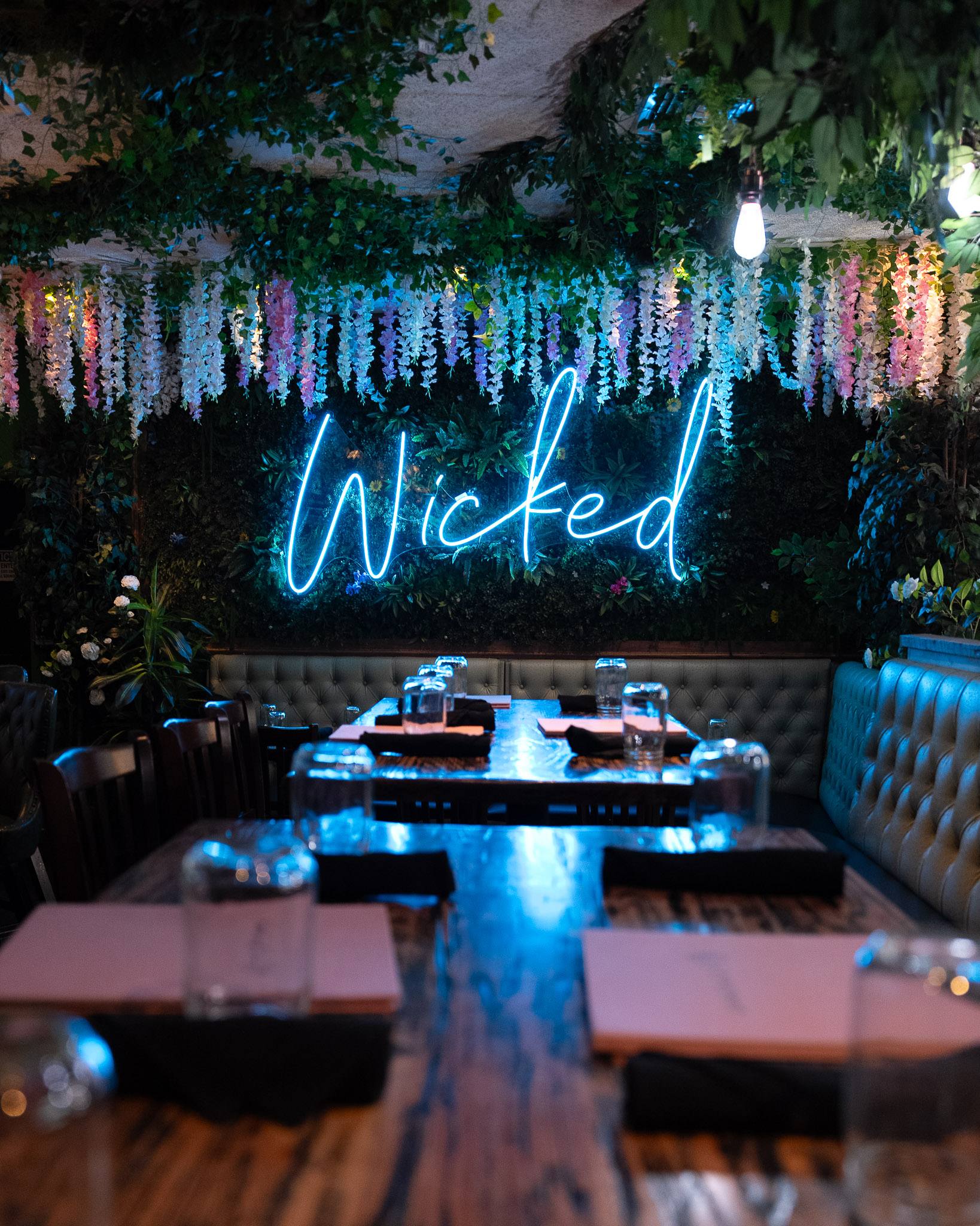 The dining room: a blue neon sign reading “Wicked” on a living green wall, pink and white wisteria hanging from the ceiling, tufted sage-green booths.