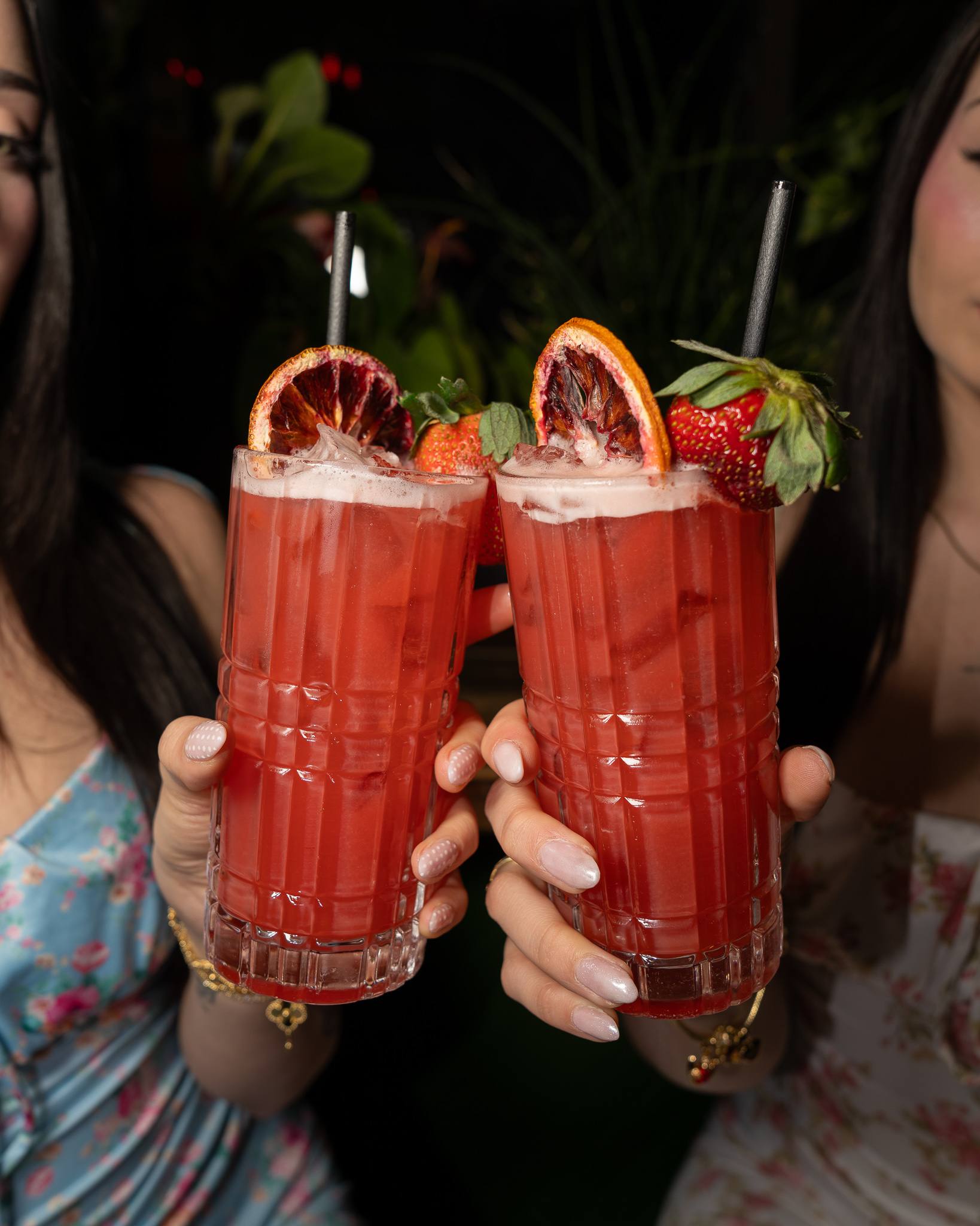 Two guests hold up tall red cocktails garnished with blood orange and whole strawberries.