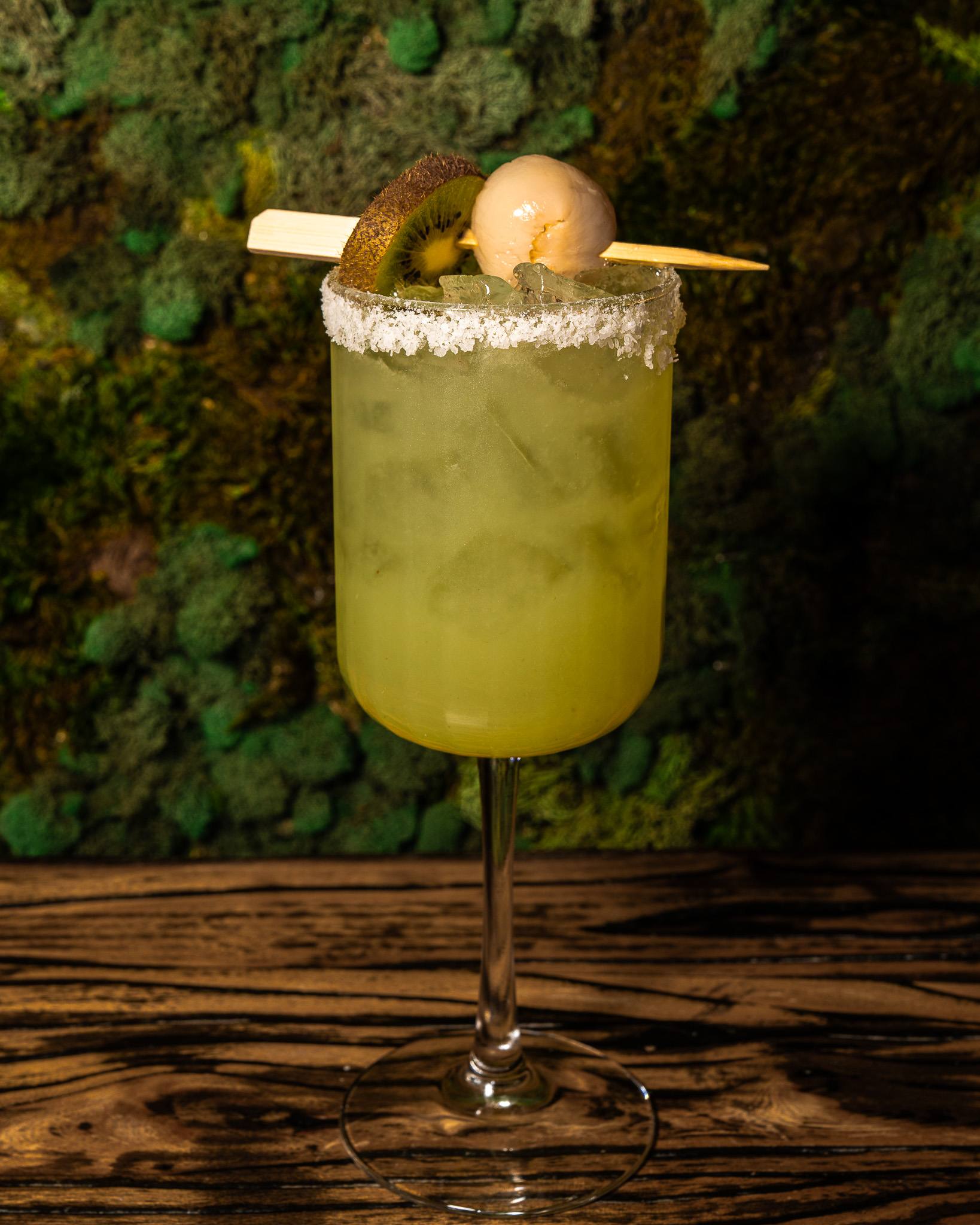 A green cocktail with a salted rim, garnished with kiwi and a lychee on a bamboo pick, against a moss wall.