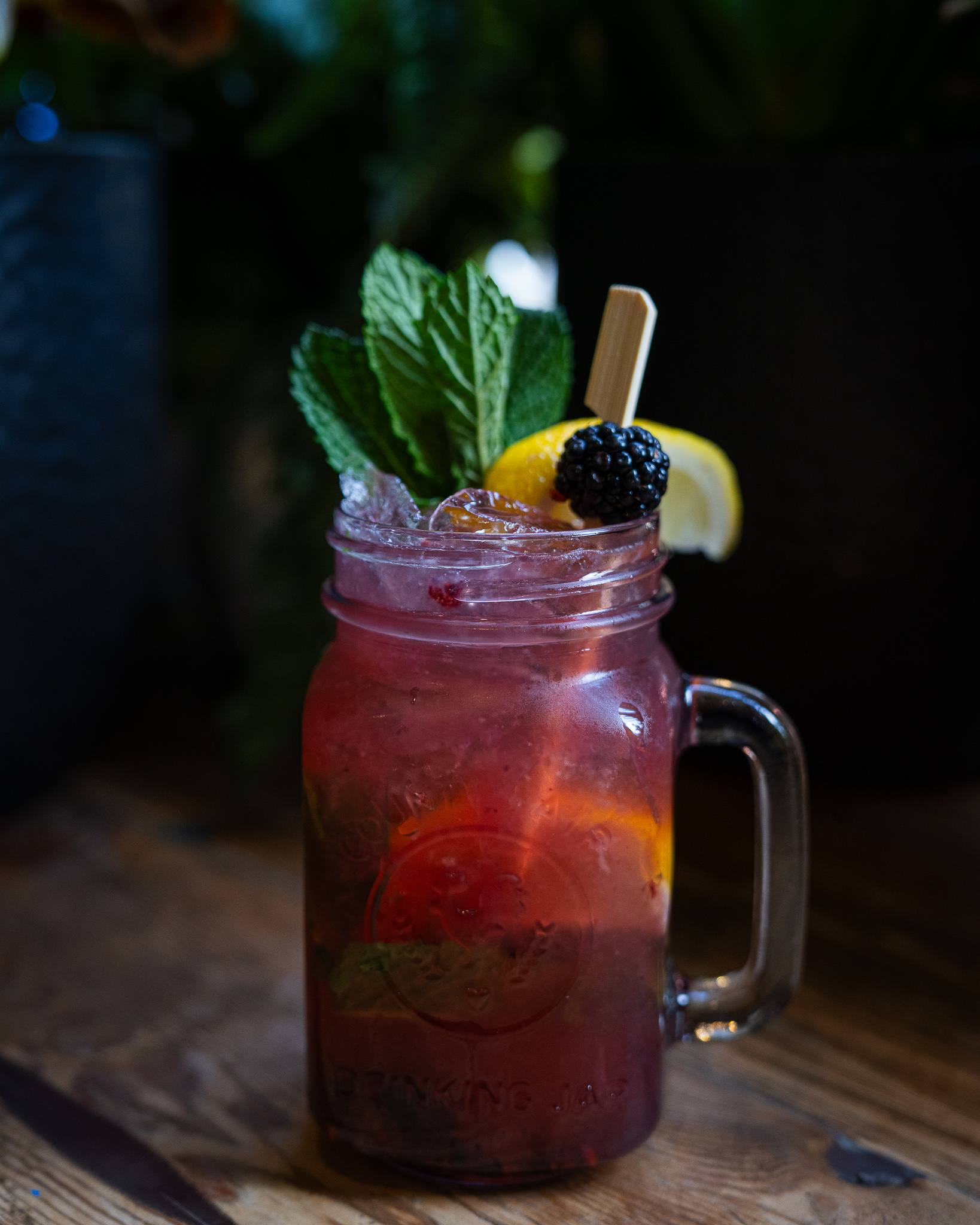 A purple-red cocktail in a mason jar mug with mint, a lemon wedge and a blackberry.