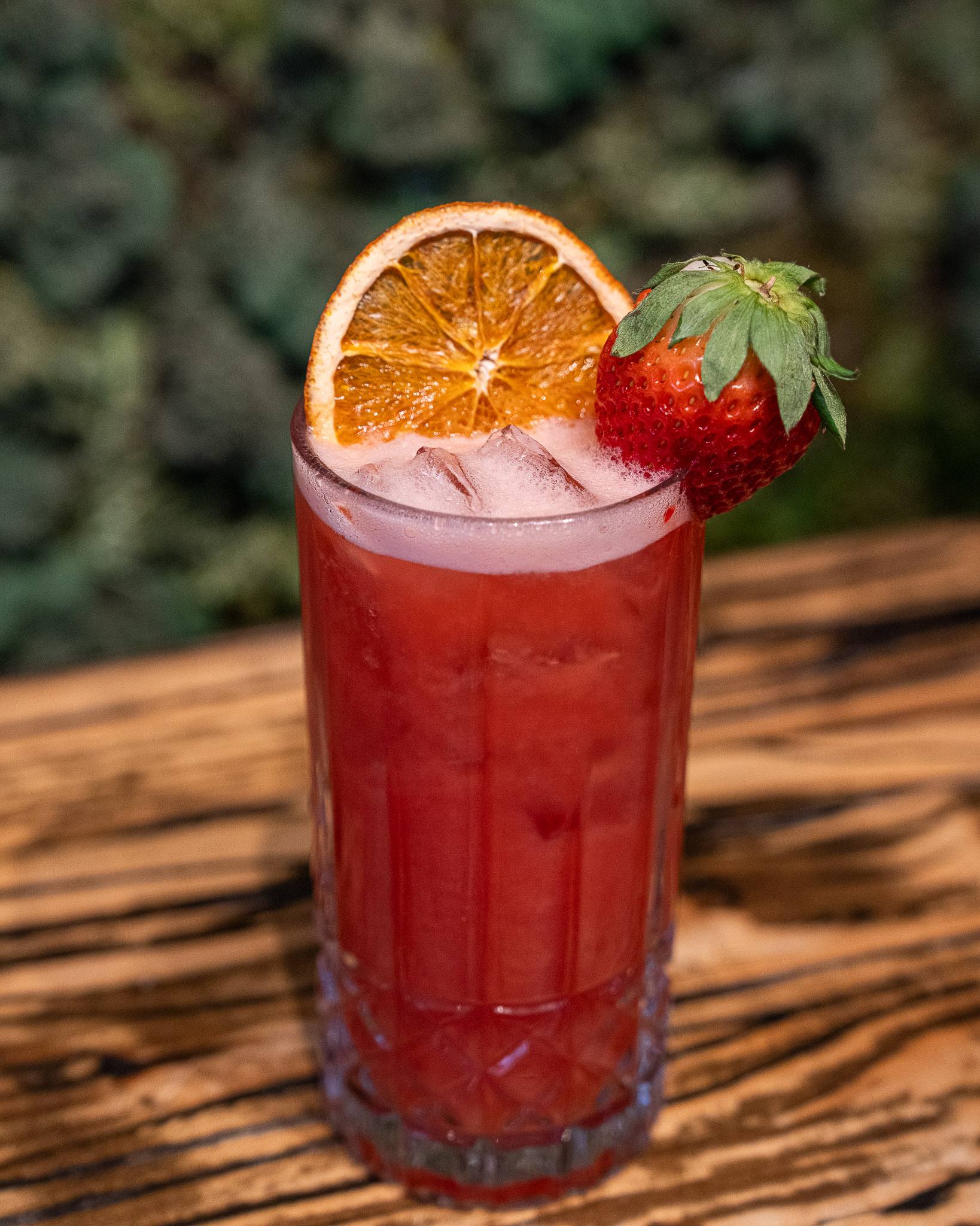 A tall strawberry-red cocktail with a dried orange wheel and a fresh strawberry on the rim.
