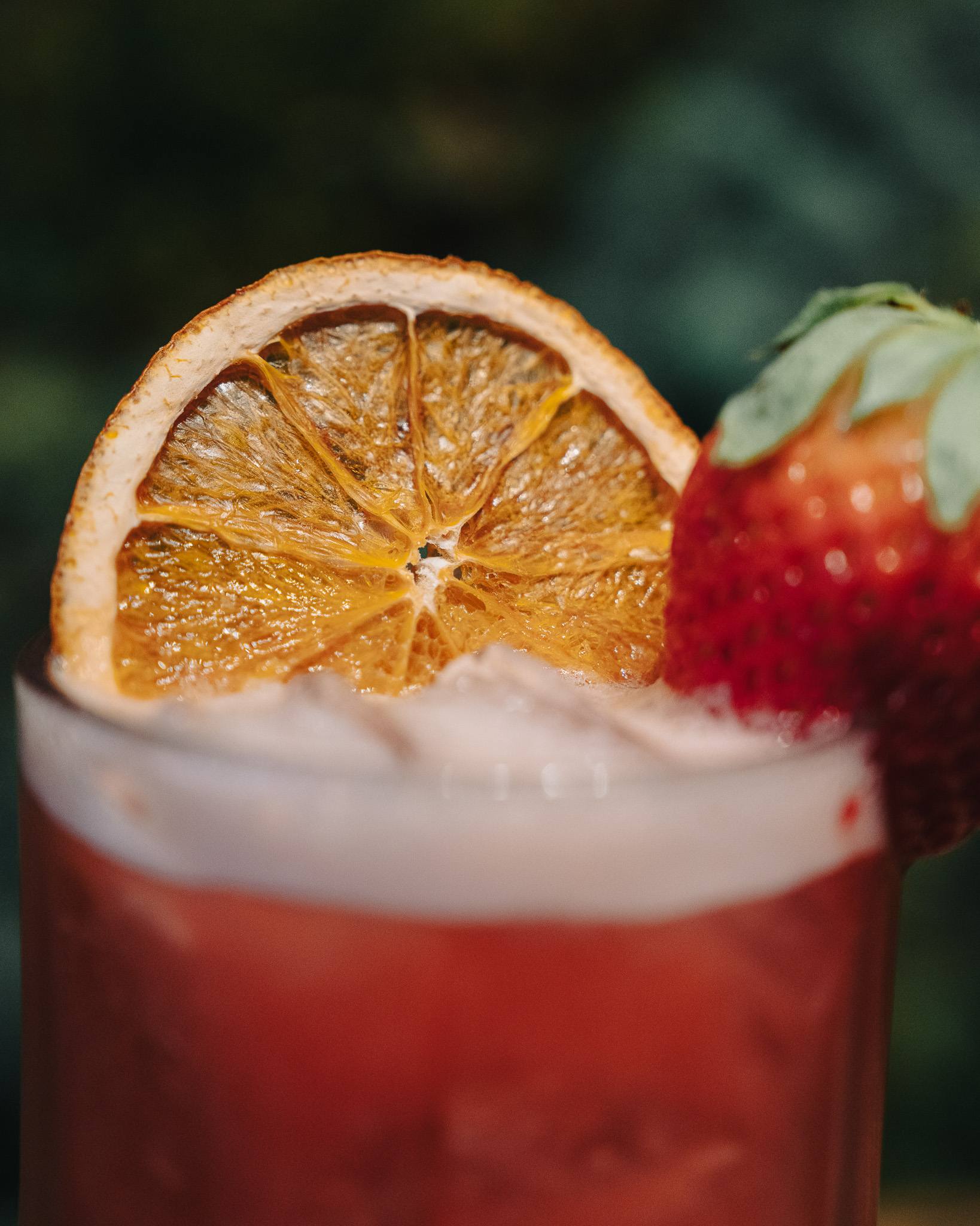 Close-up of a dried orange wheel and a strawberry on the rim of a red cocktail.