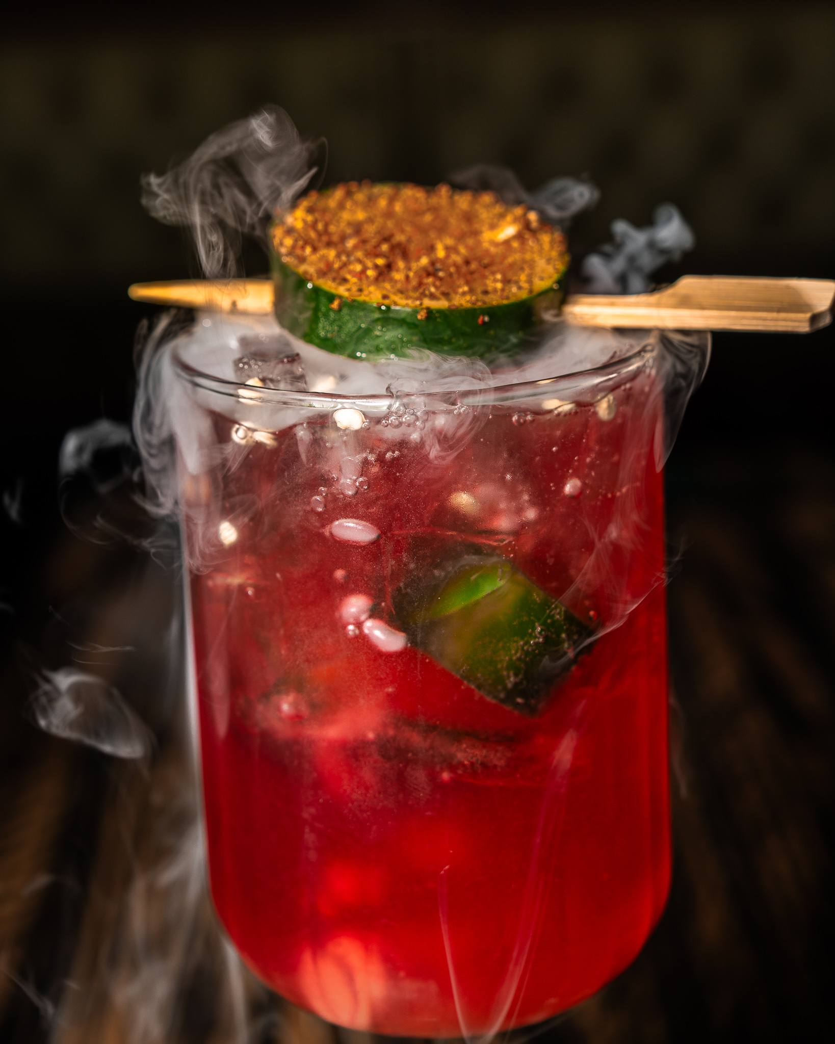 A bright red cocktail wrapped in dry-ice smoke, topped with a chili-dusted cucumber wheel on a bamboo pick.