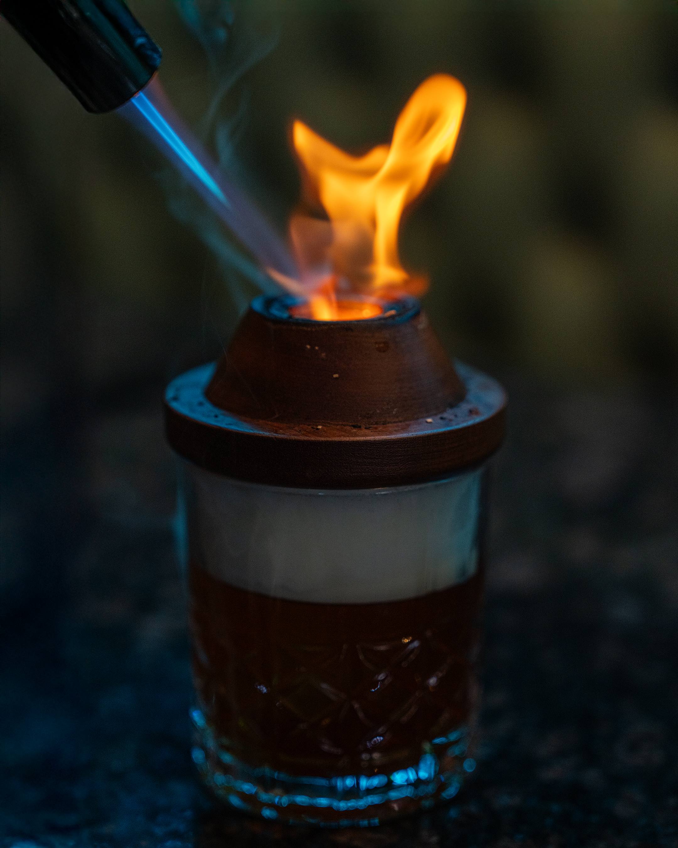 A bartender’s torch lights a wooden smoker on top of a cut-crystal rocks glass, flame rising.