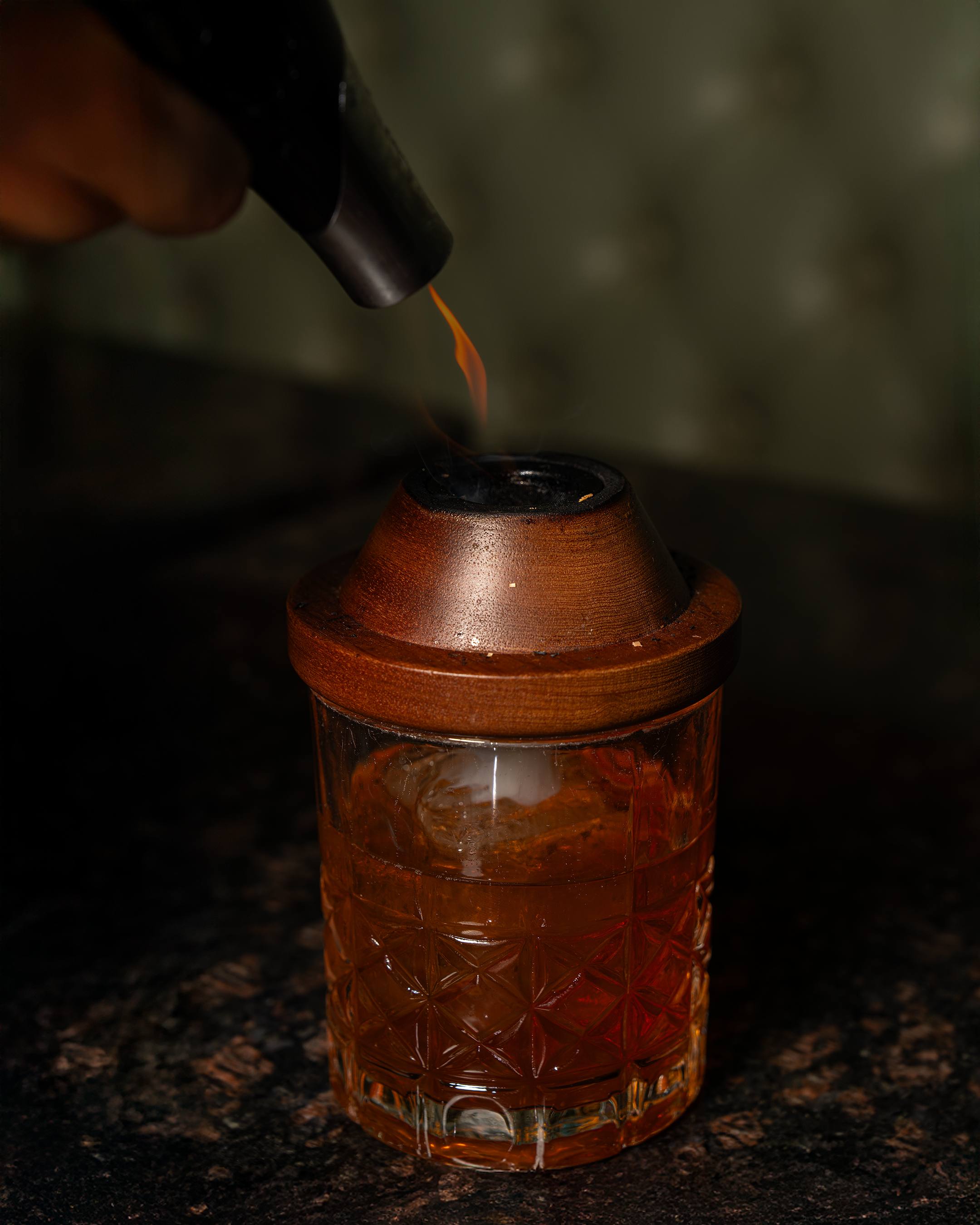 A torch is lowered toward a wooden smoker sitting on a crystal rocks glass.