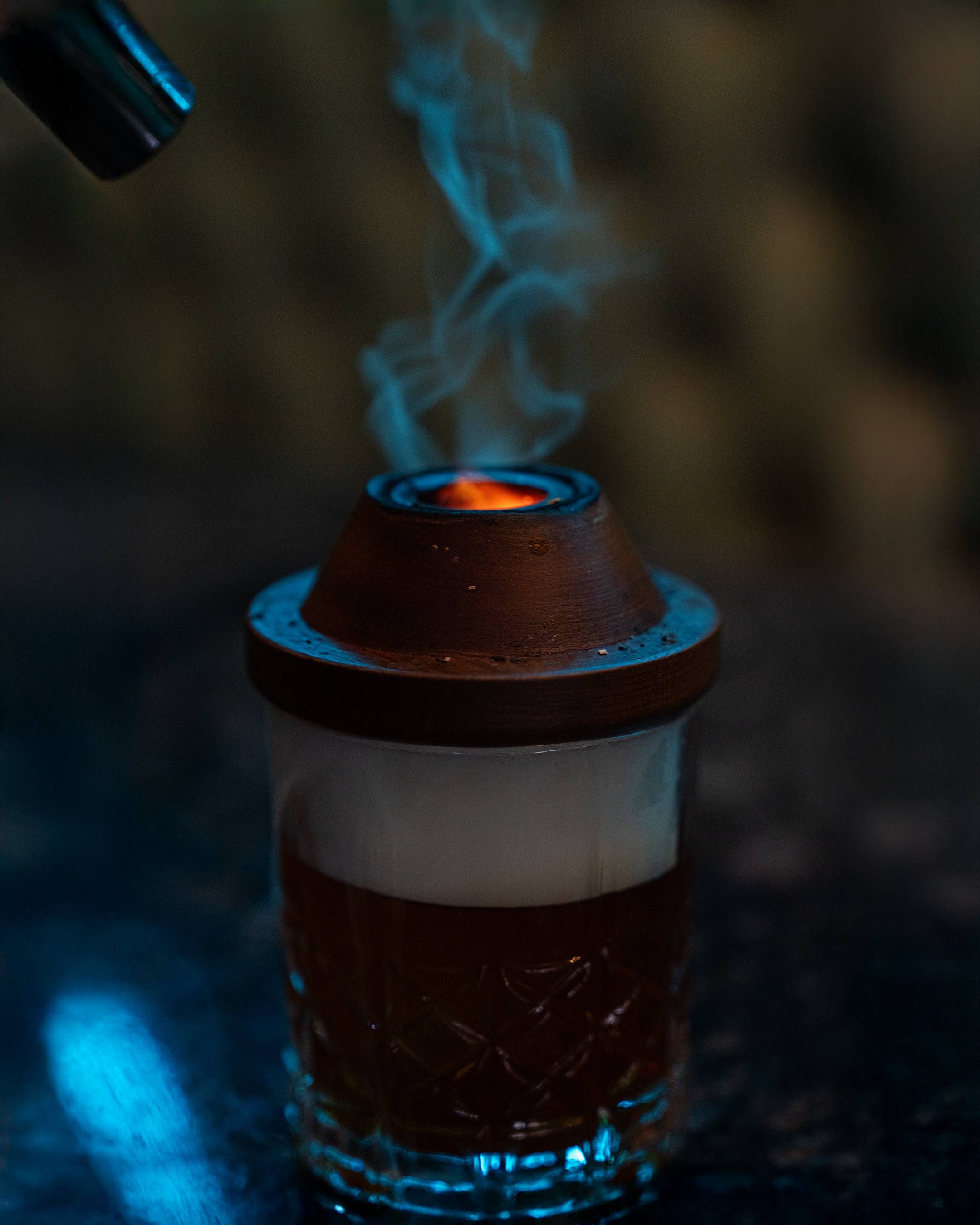 Blue-lit smoke rises from the wooden smoker on top of an old fashioned.