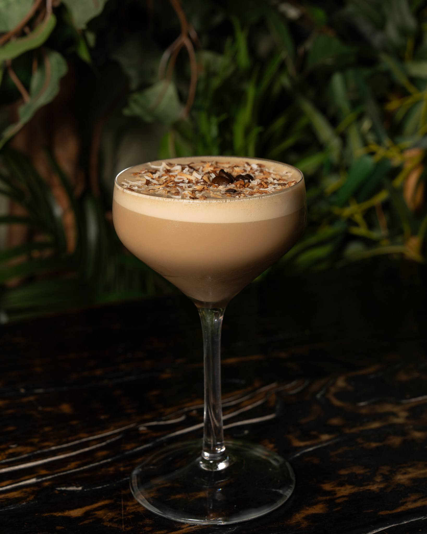 An espresso martini in a coupe topped with toasted coconut and three coffee beans, in front of tropical plants.