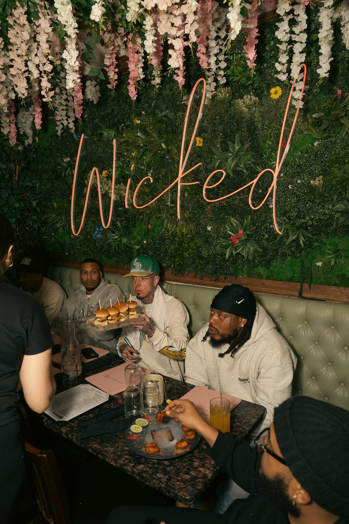 A group of friends in a booth beneath the neon sign, glowing coral, one holding up a board of crispy chicken sliders.