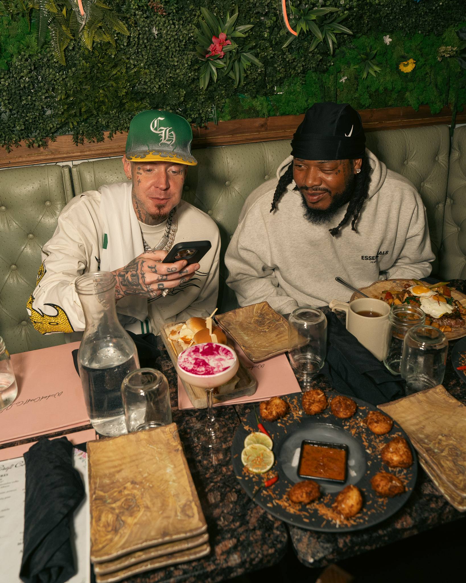Two guests laughing in a sage-green booth, a plate of coconut shrimp and a cocktail with printed foam on the table.