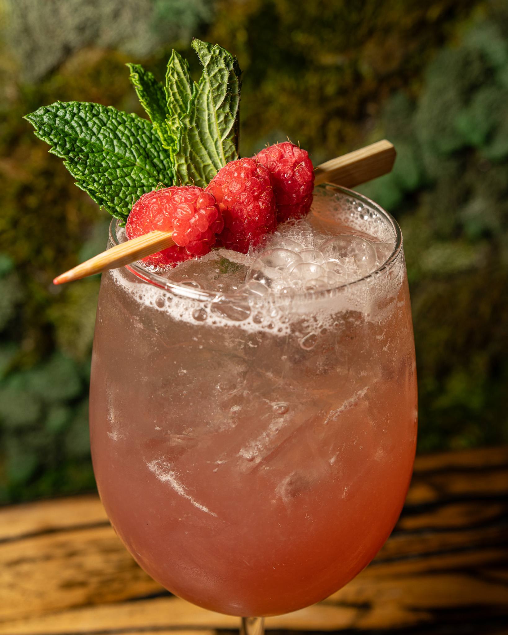 A pink spritz over crushed ice, garnished with a skewer of raspberries and a sprig of mint, against the moss wall.