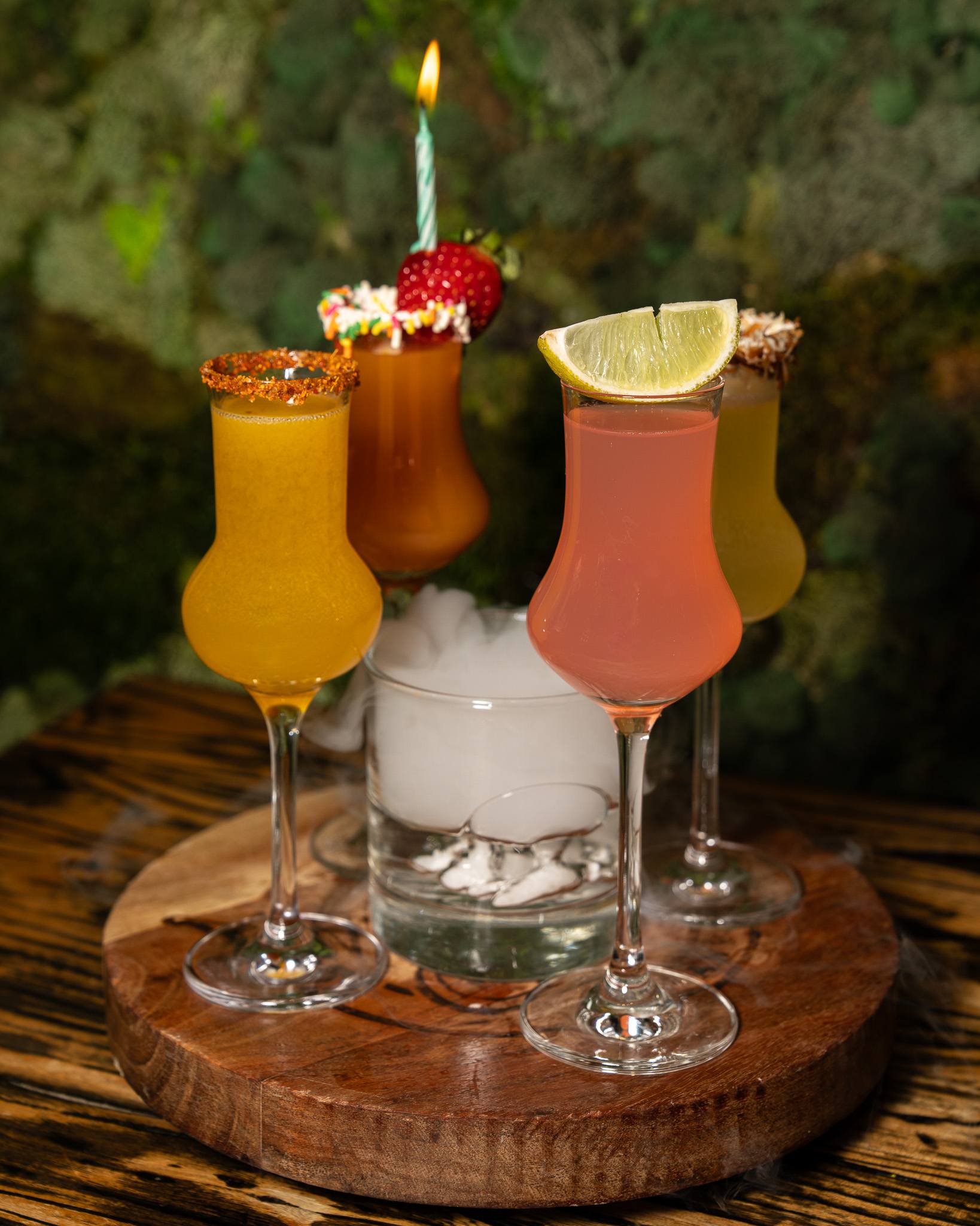 A flight of four specialty shots in tulip glasses around a glass of dry ice on a wooden board, one with a sprinkle rim, a strawberry and a lit birthday candle.