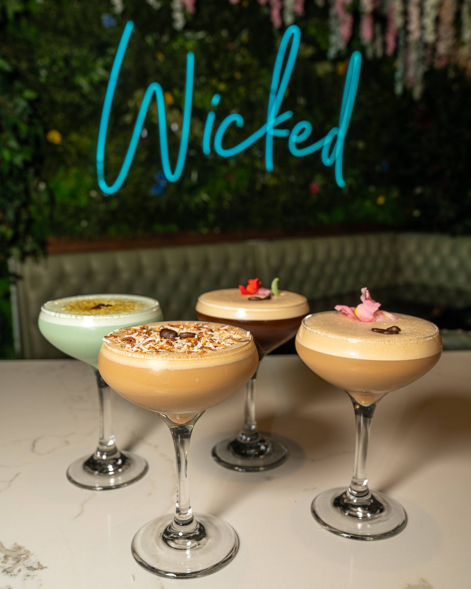 Four espresso martinis in coupes on a marble bar, one pistachio green, one topped with toasted coconut, two with flower petals, the blue neon “Wicked” sign behind them.