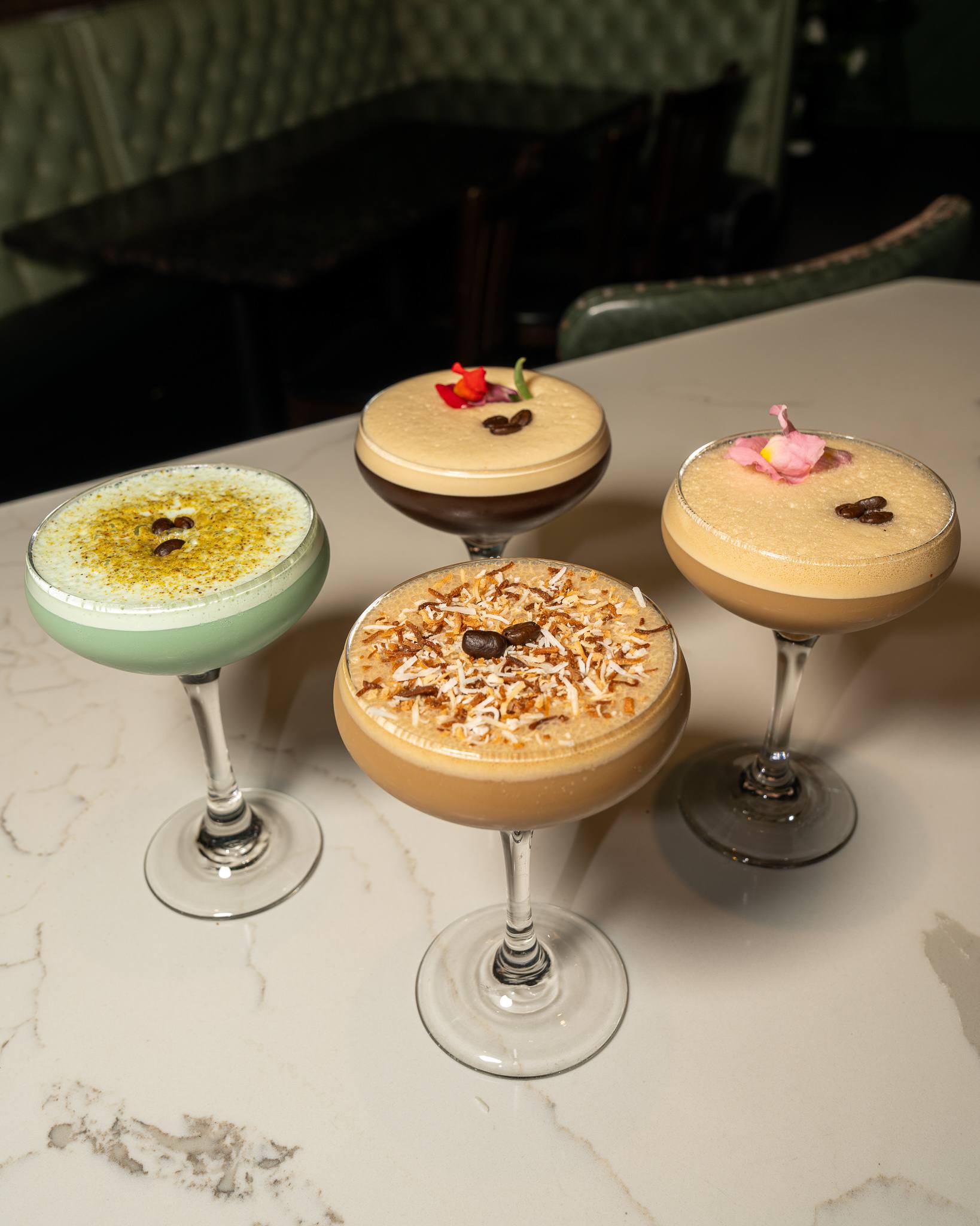 Four espresso martinis seen from above on a marble bar: pistachio green, toasted coconut, and two with flower petals and coffee beans.