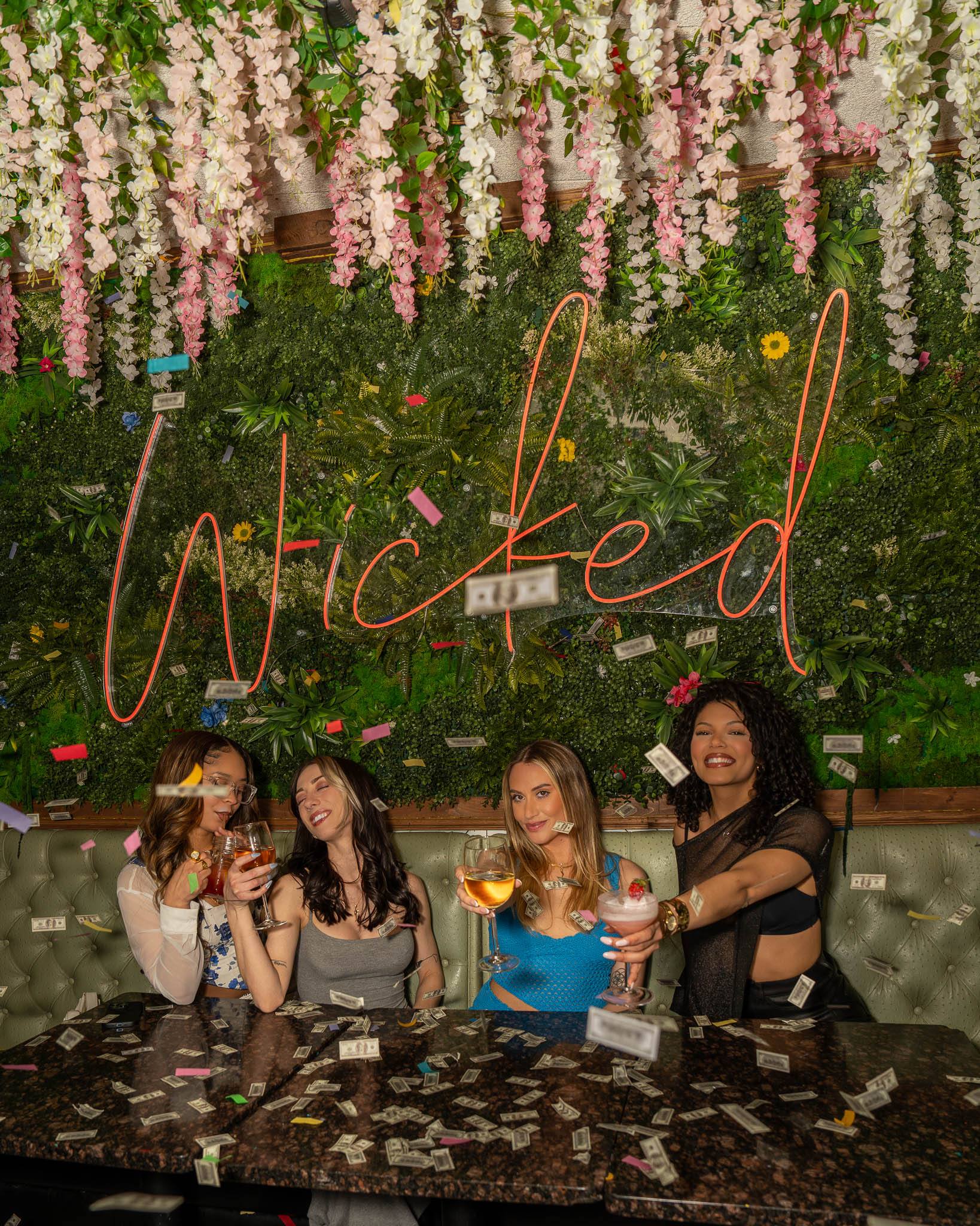 Four friends raise their glasses in a sage-green booth as confetti and play money rain down, the neon sign glowing coral on the flower wall above them.