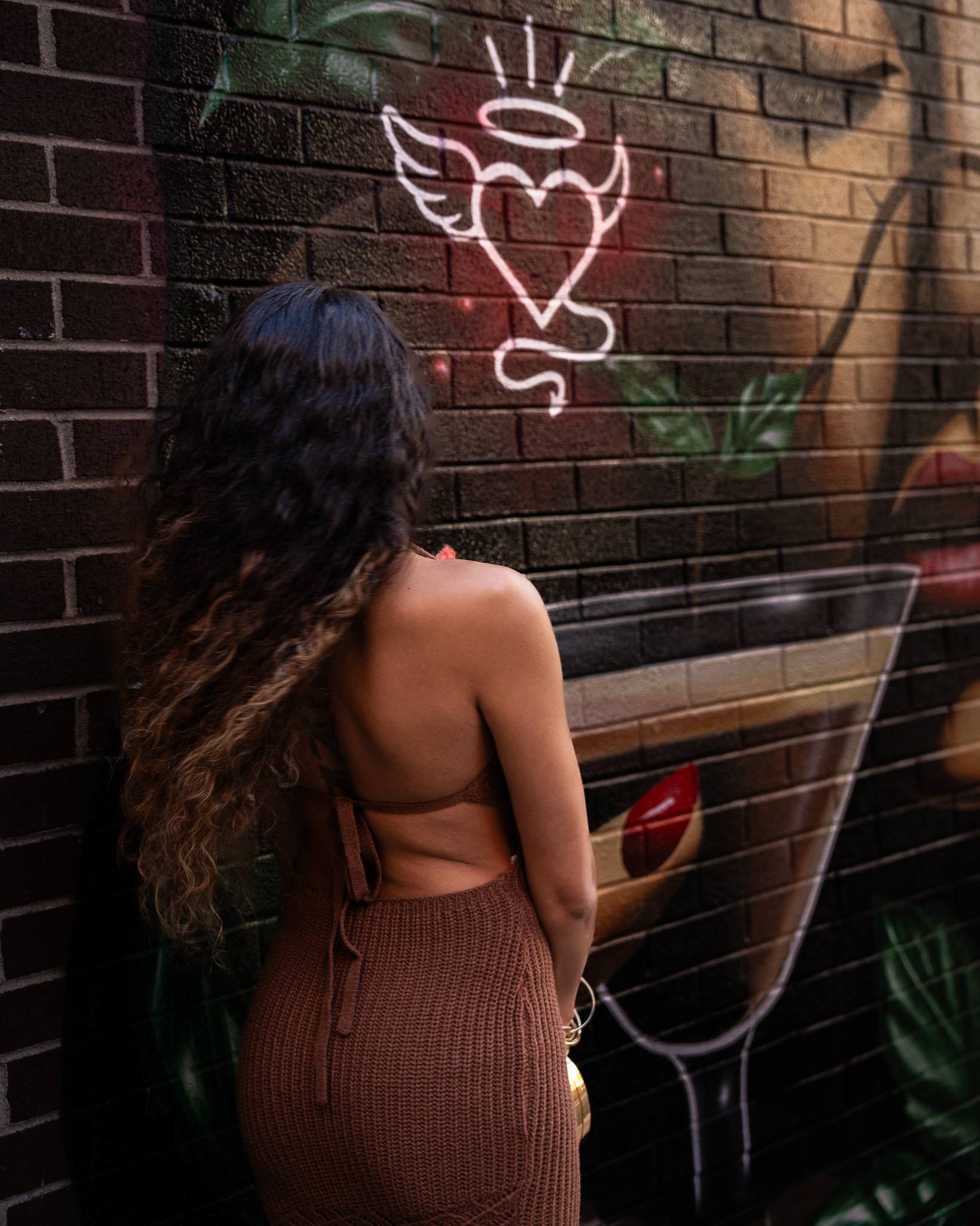 A guest with long curls stands with her back to the camera at the mural outside, beside a painted winged heart and a martini glass.