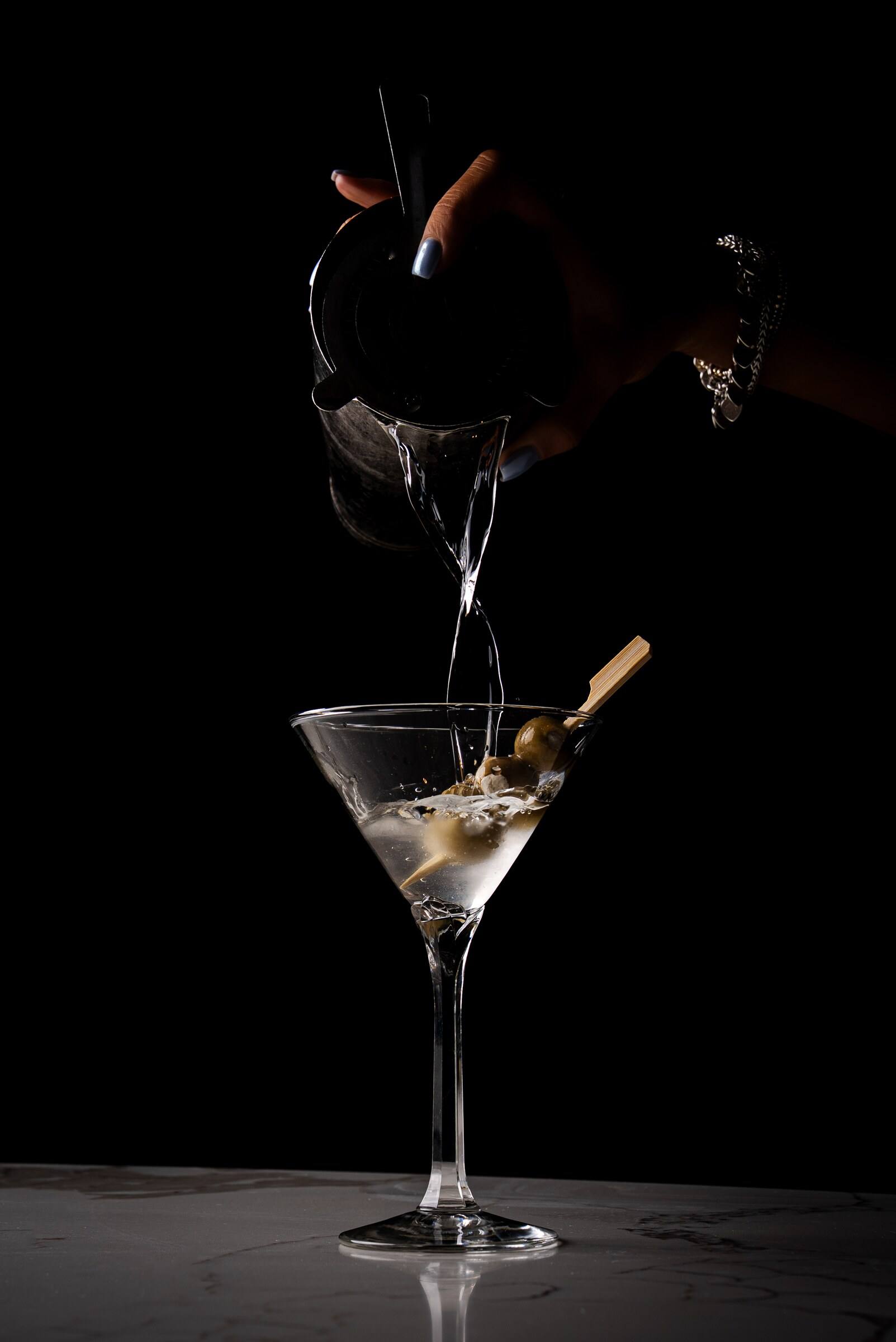 A bartender pours a clear martini into an olive-garnished glass against a black background.