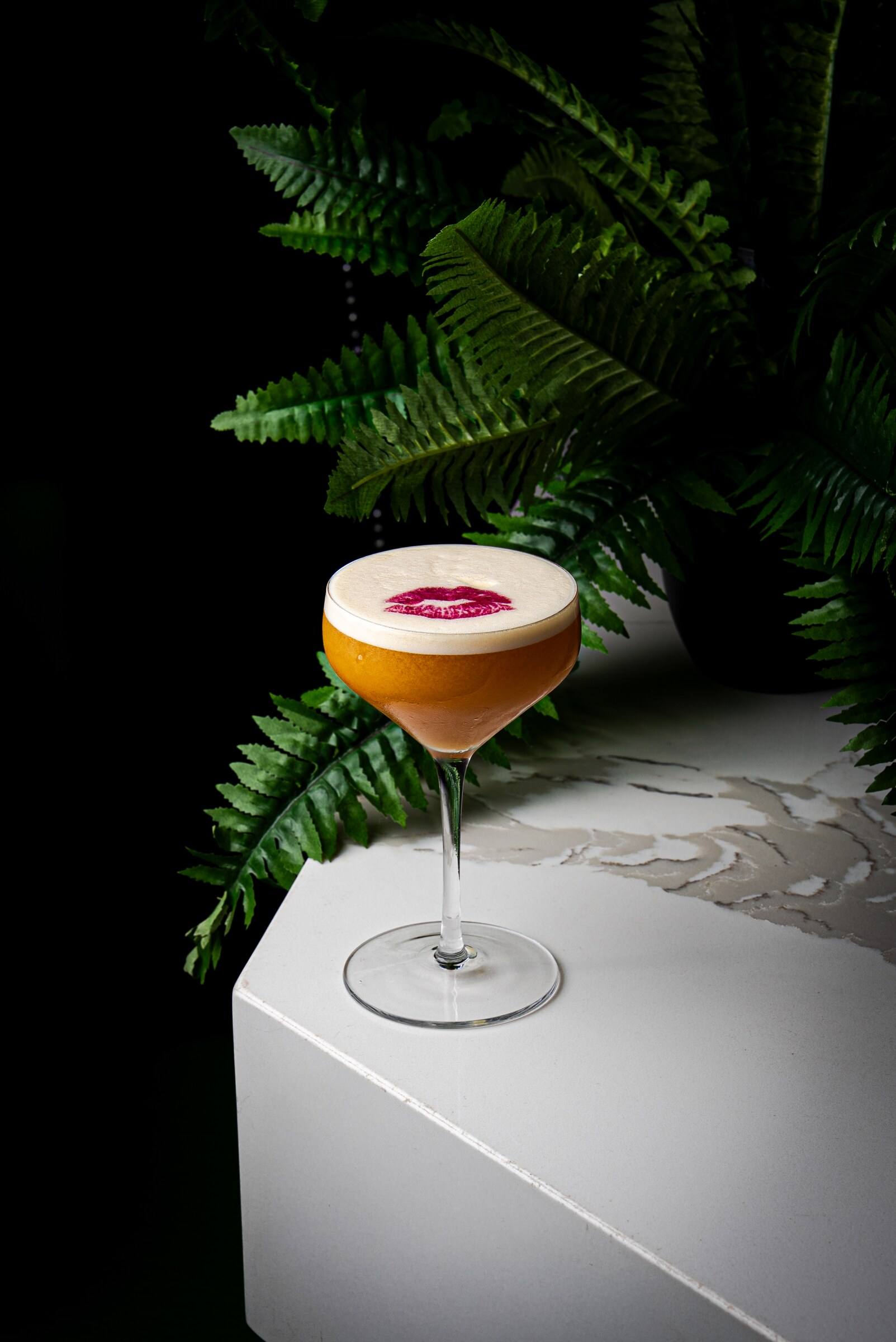 An amber cocktail in a coupe with a pink lipstick kiss printed across the foam.