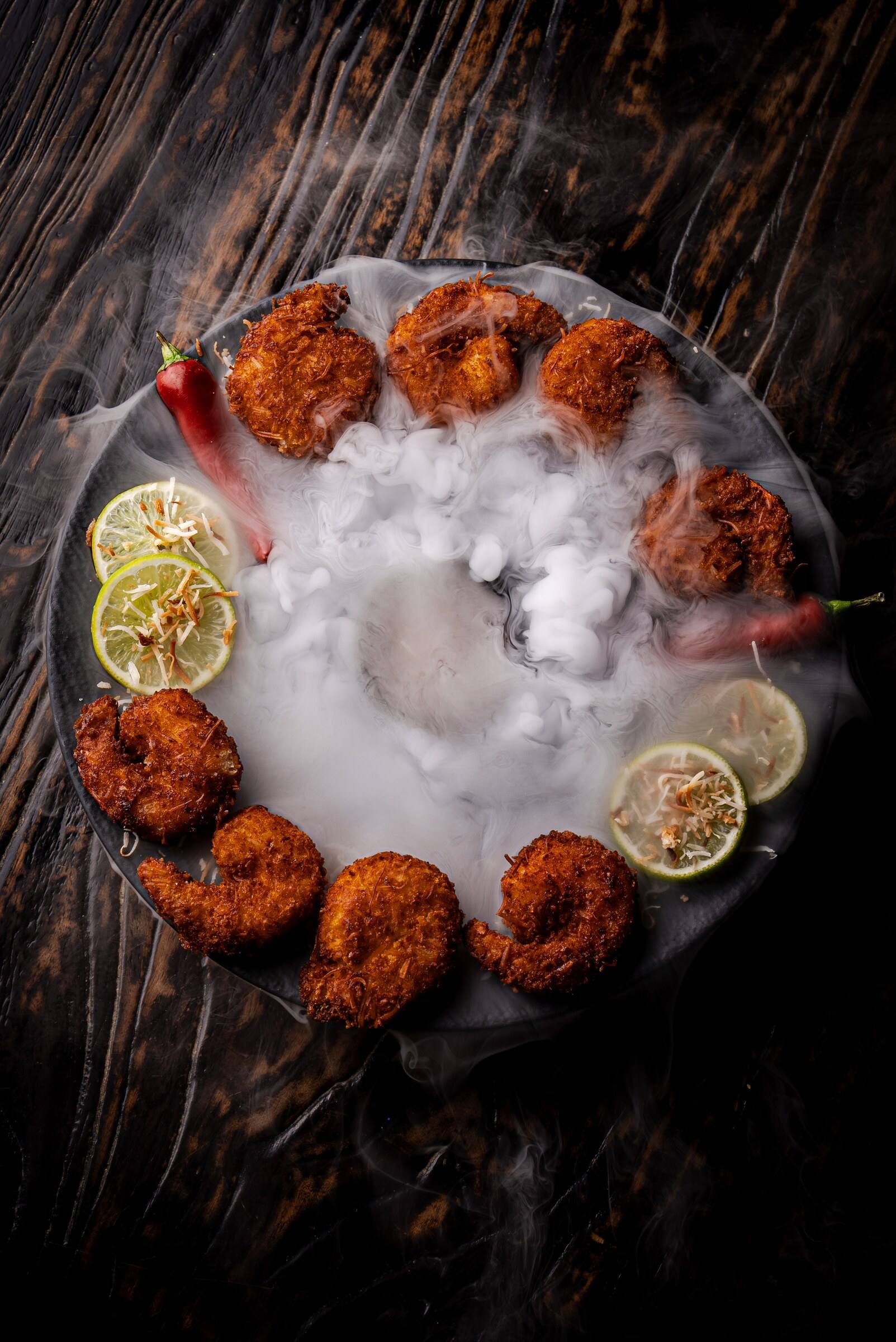 Coconut-crusted shrimp arranged around a smoking platter with lime slices and red peppers.