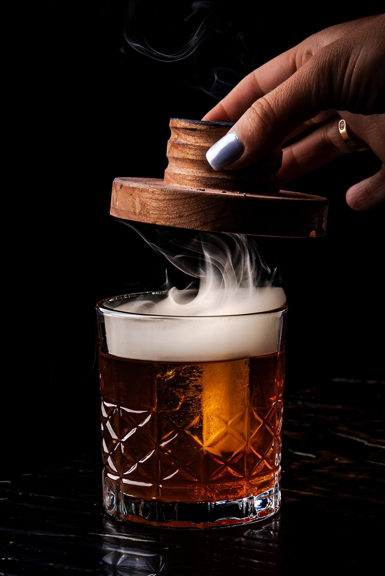 A bartender lifts a wooden smoker from a cut-crystal rocks glass as white smoke rolls over an amber cocktail.