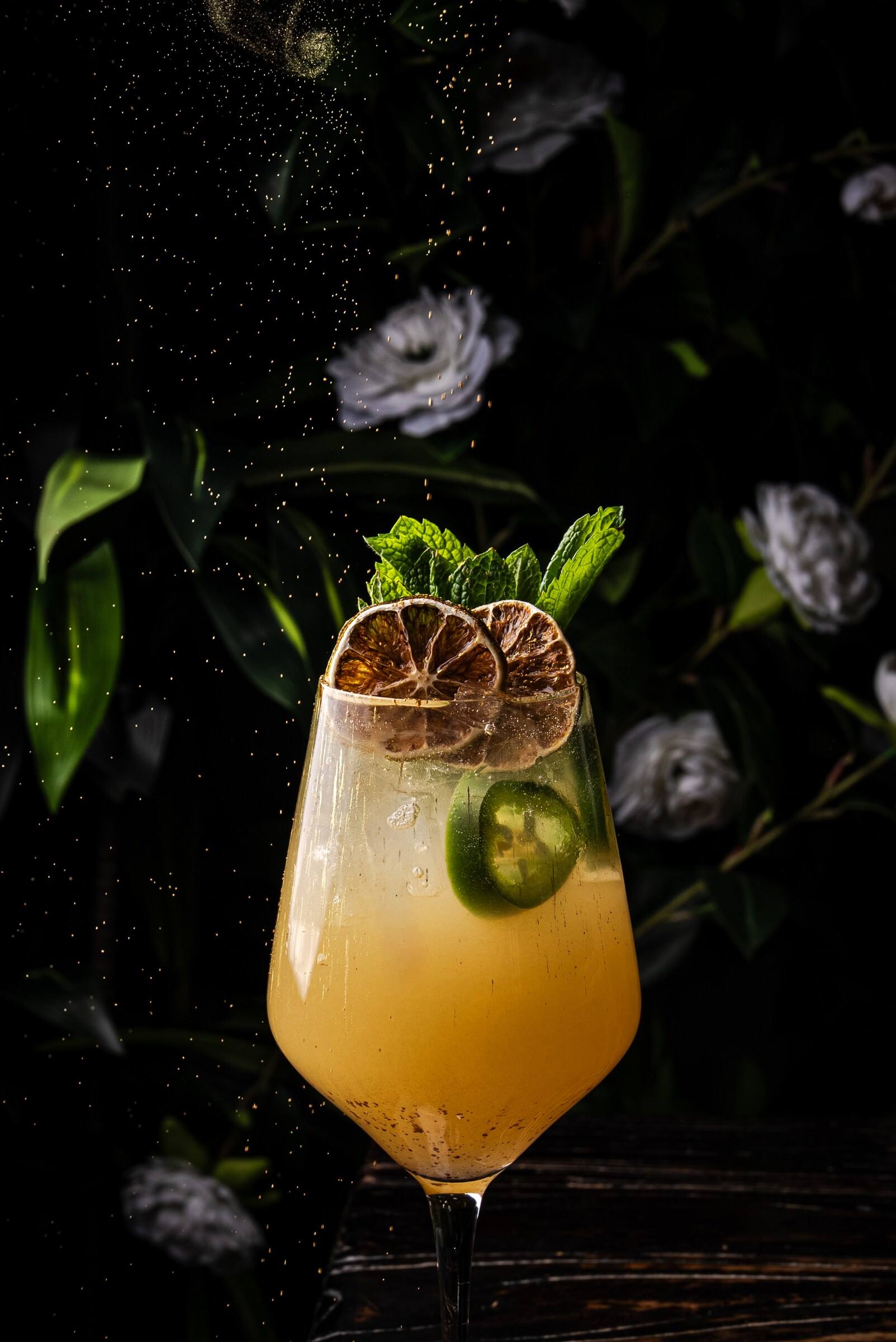 A golden cocktail with jalapeño, dried citrus and mint as gold flecks fall against the flower wall.