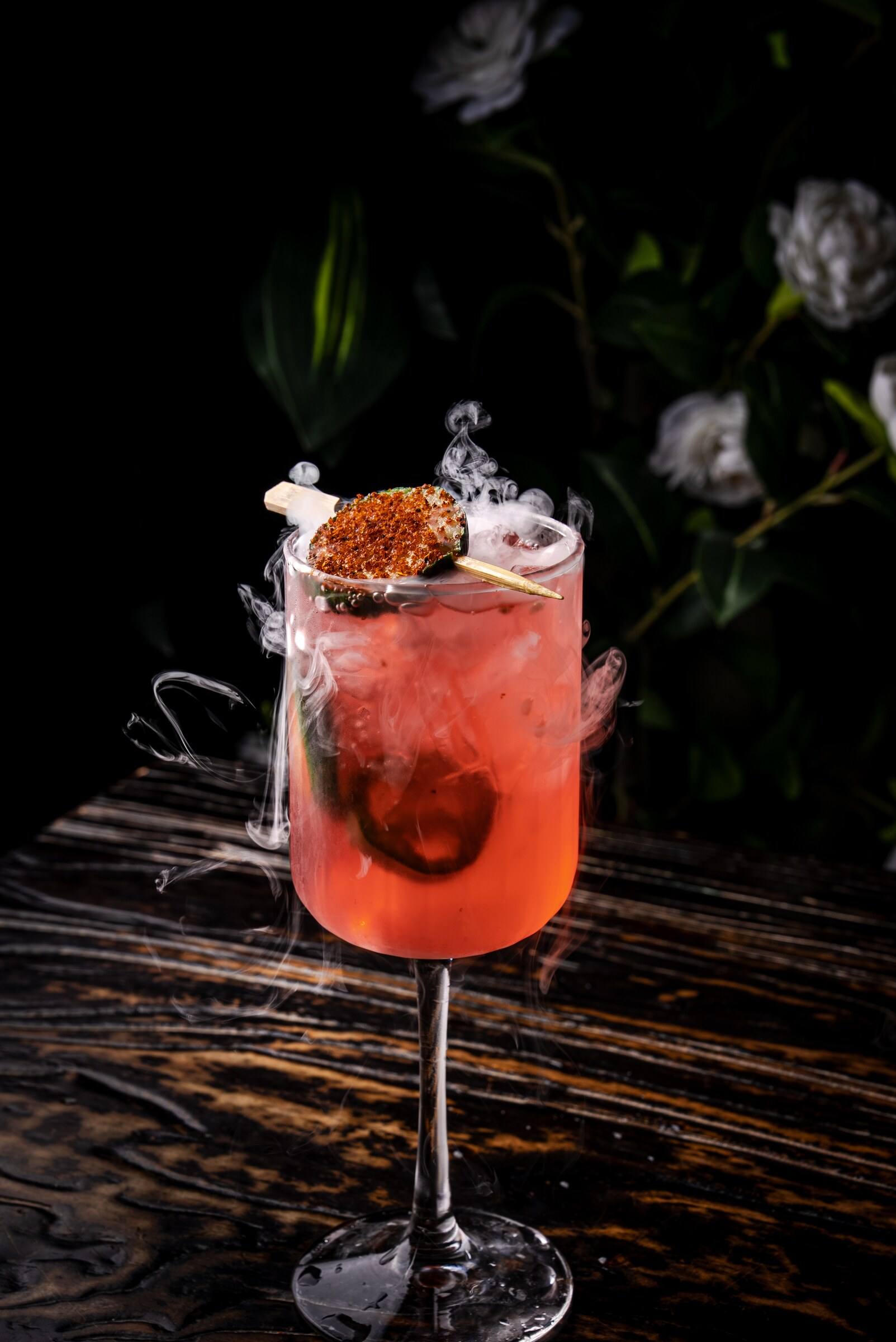 A bright pink cocktail in a stemmed glass surrounded by curling white smoke.