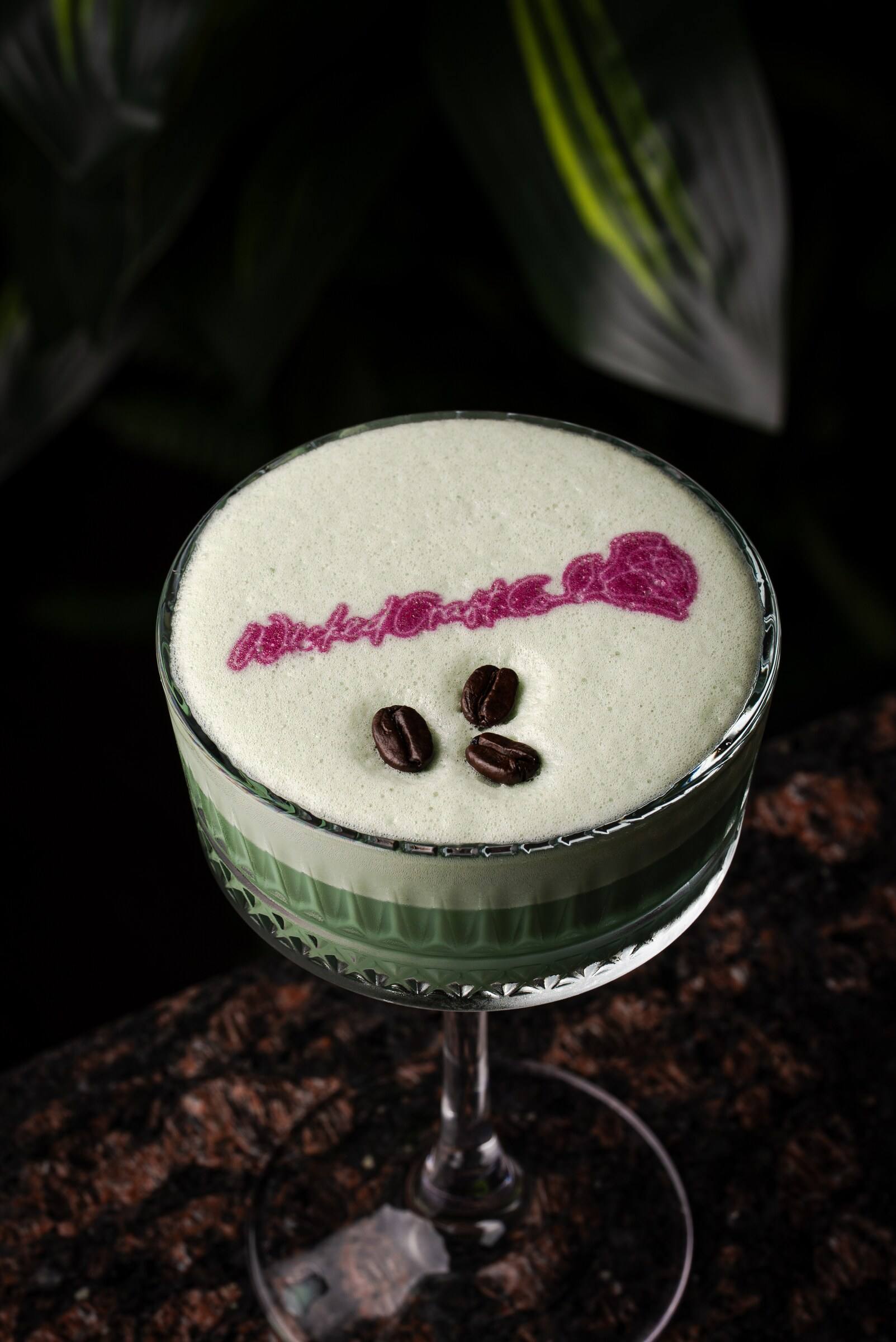 A pale green foam-topped cocktail printed with a pink message and garnished with coffee beans.