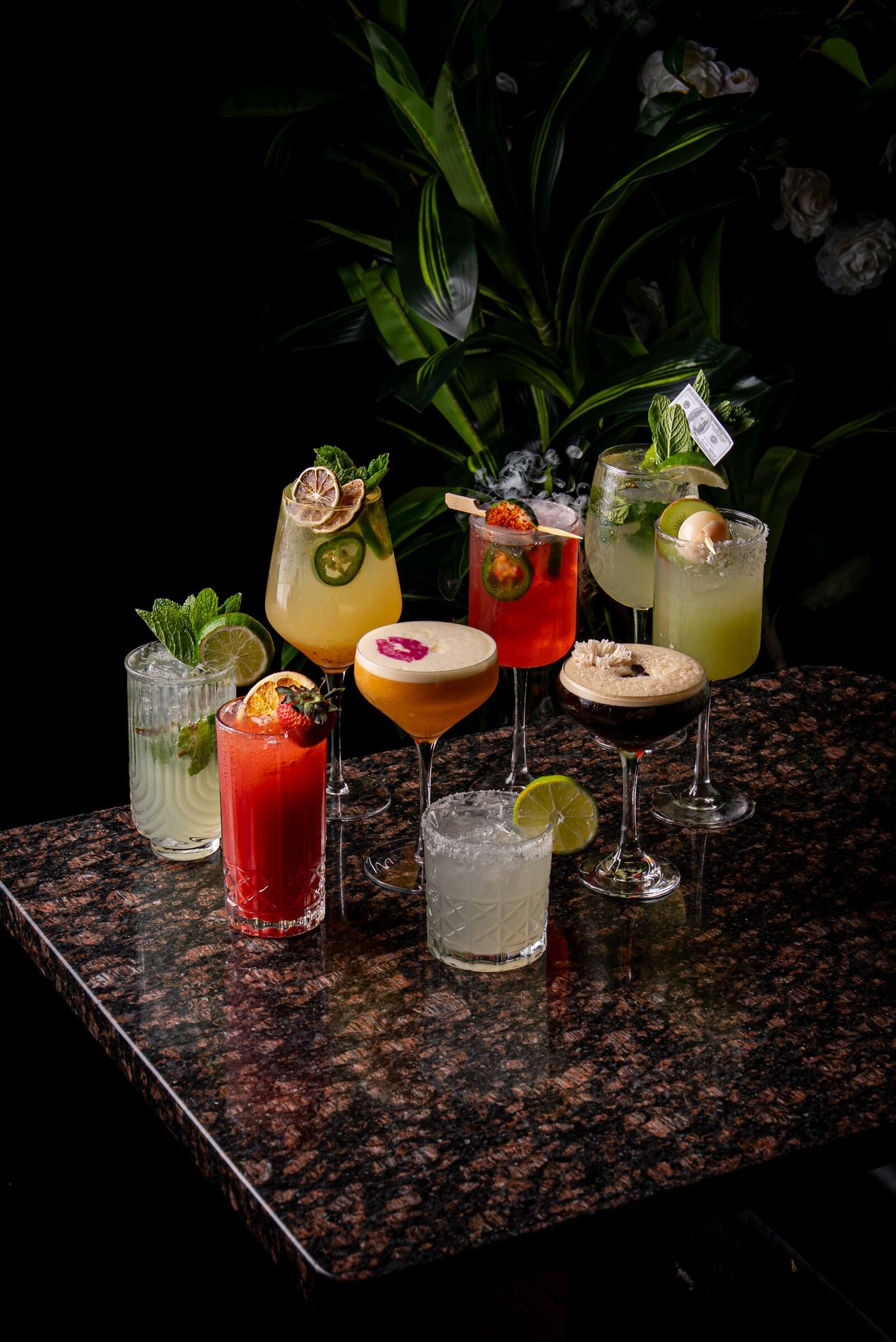 Nine colorful cocktails arranged across a granite table beneath tropical plants.