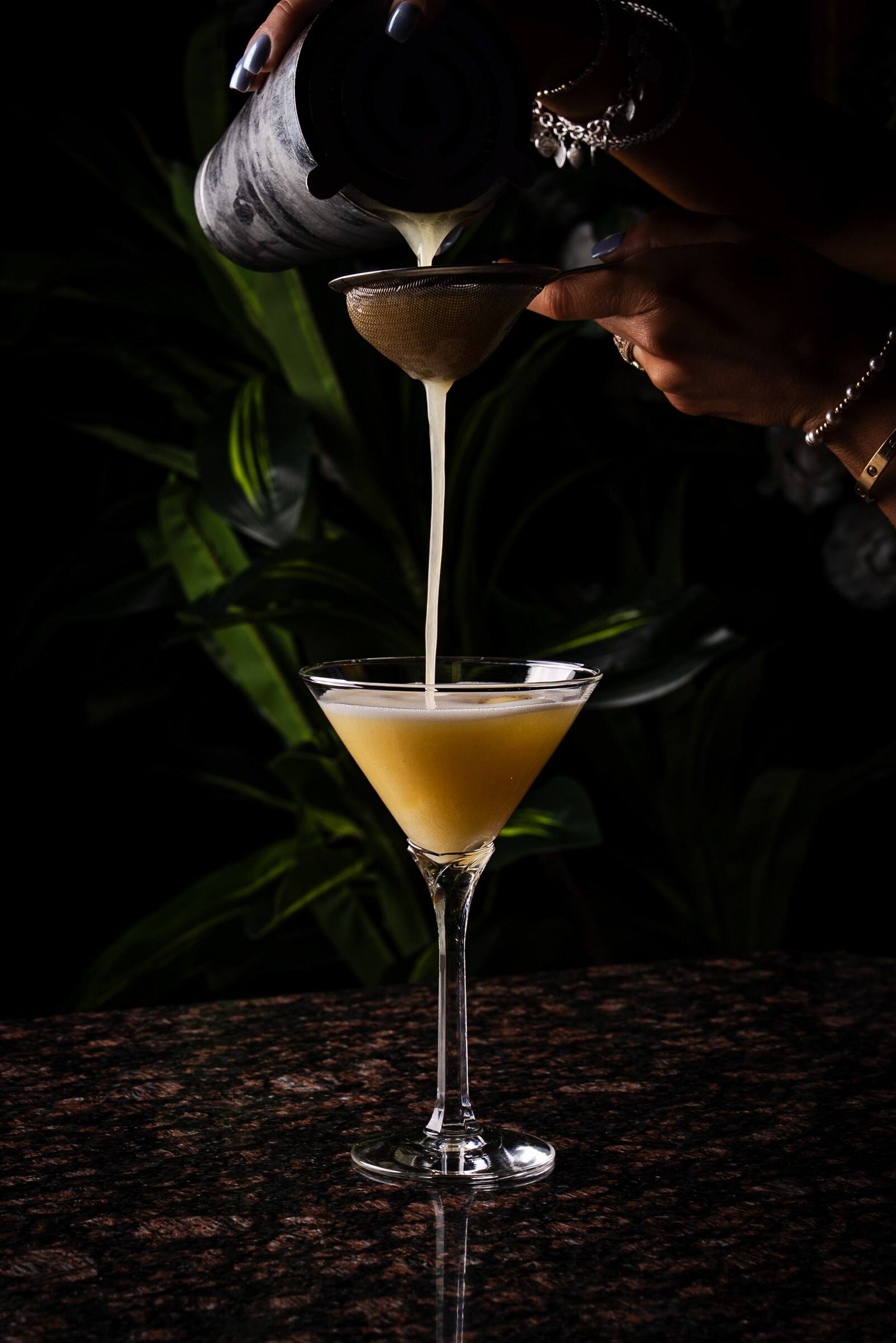 A bartender double-strains a golden cocktail from a shaker into a martini glass.