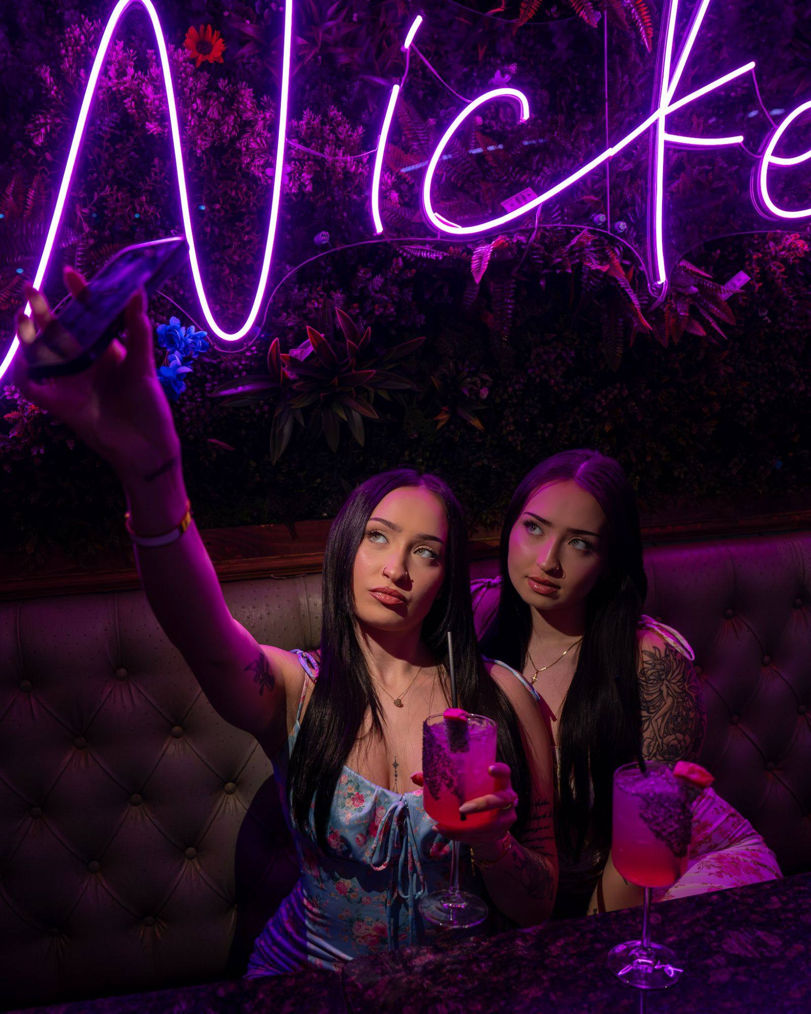 Two friends in a tufted booth beneath the “Wicked” neon glowing purple on the flower wall, one holding her phone up for a selfie, both with pink cocktails in stemmed glasses.
