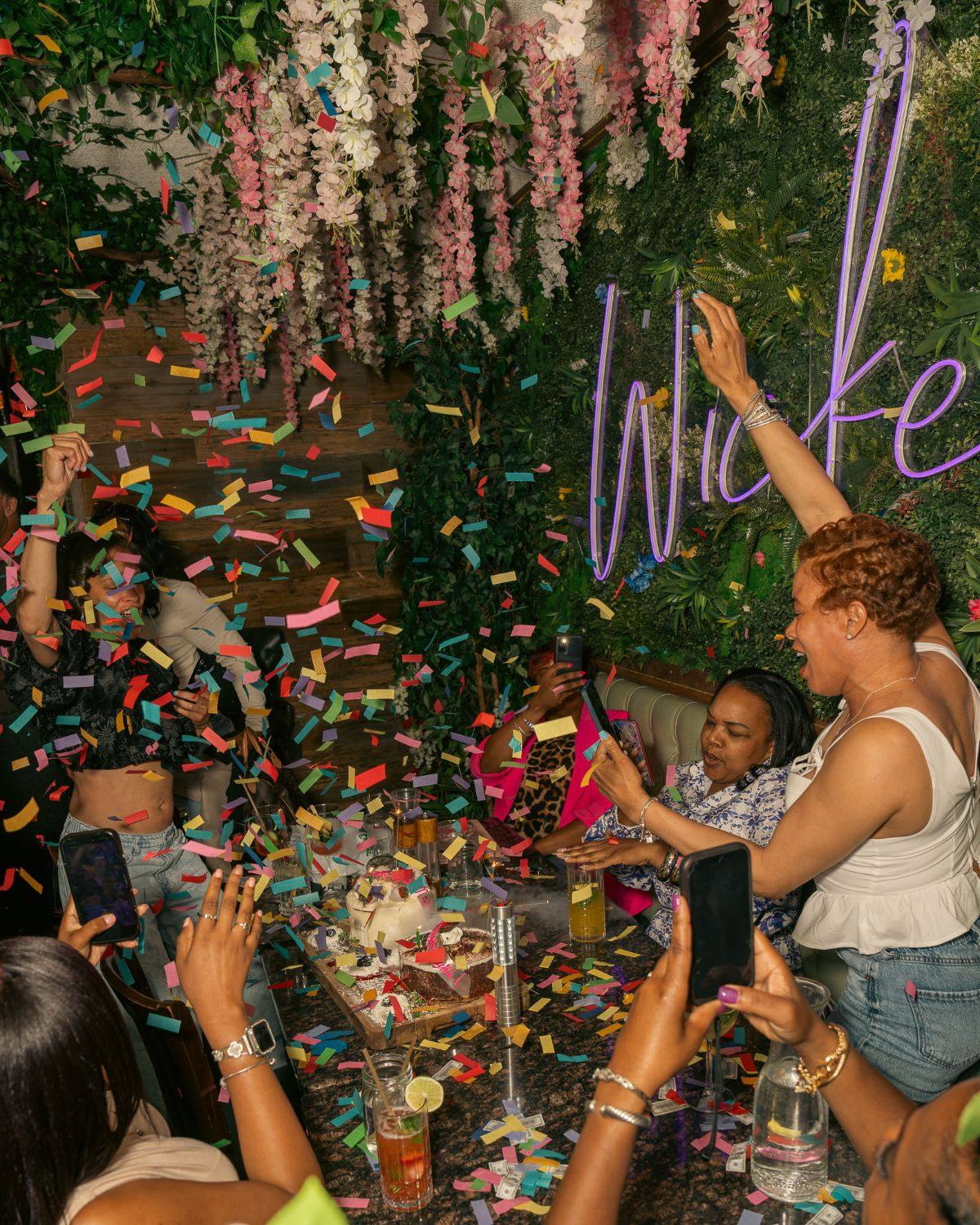 Confetti rains over a table of friends celebrating under hanging wisteria and the purple “Wicked” neon, arms in the air, phones out, a cake and desserts on the table.