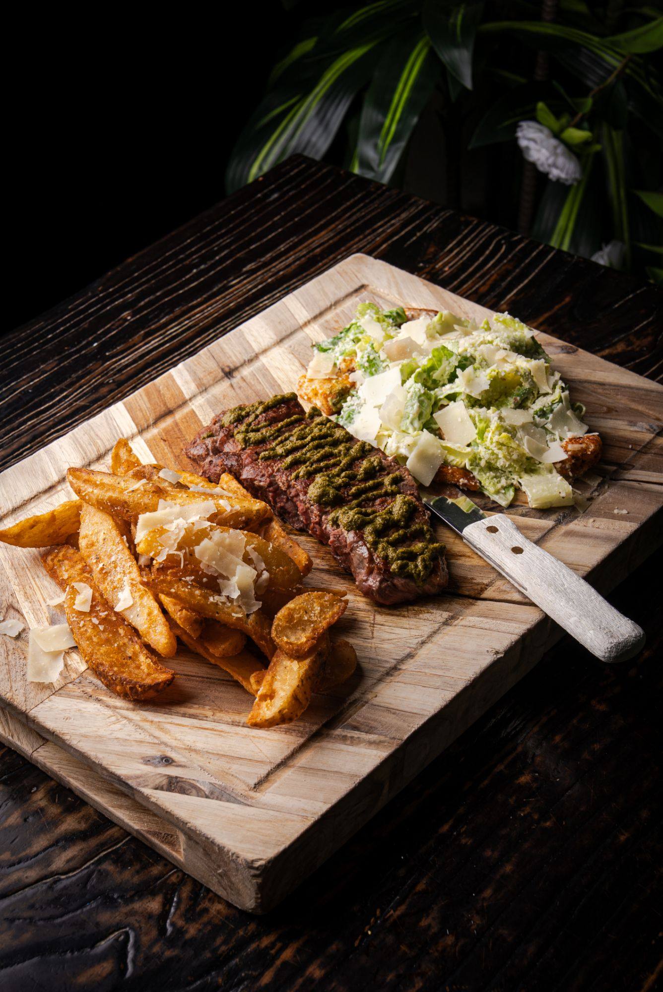 Sliced grilled sirloin drizzled with green chimichurri on a wooden board, with Parmesan-topped wedge fries, a Caesar salad and a steak knife.