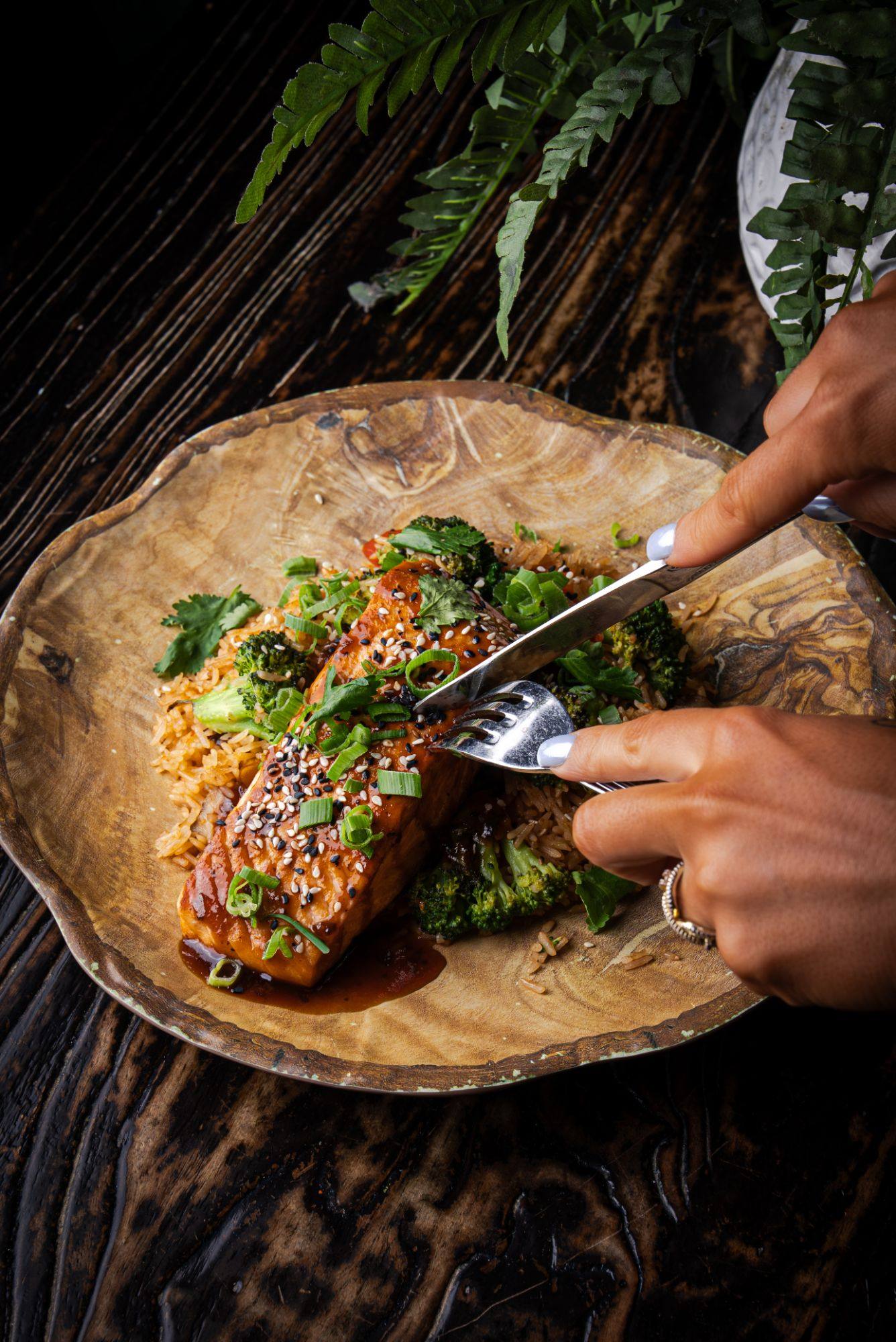 A guest cuts into a teriyaki-glazed salmon fillet topped with sesame seeds, scallions and cilantro, over rice and broccoli on a wooden plate.